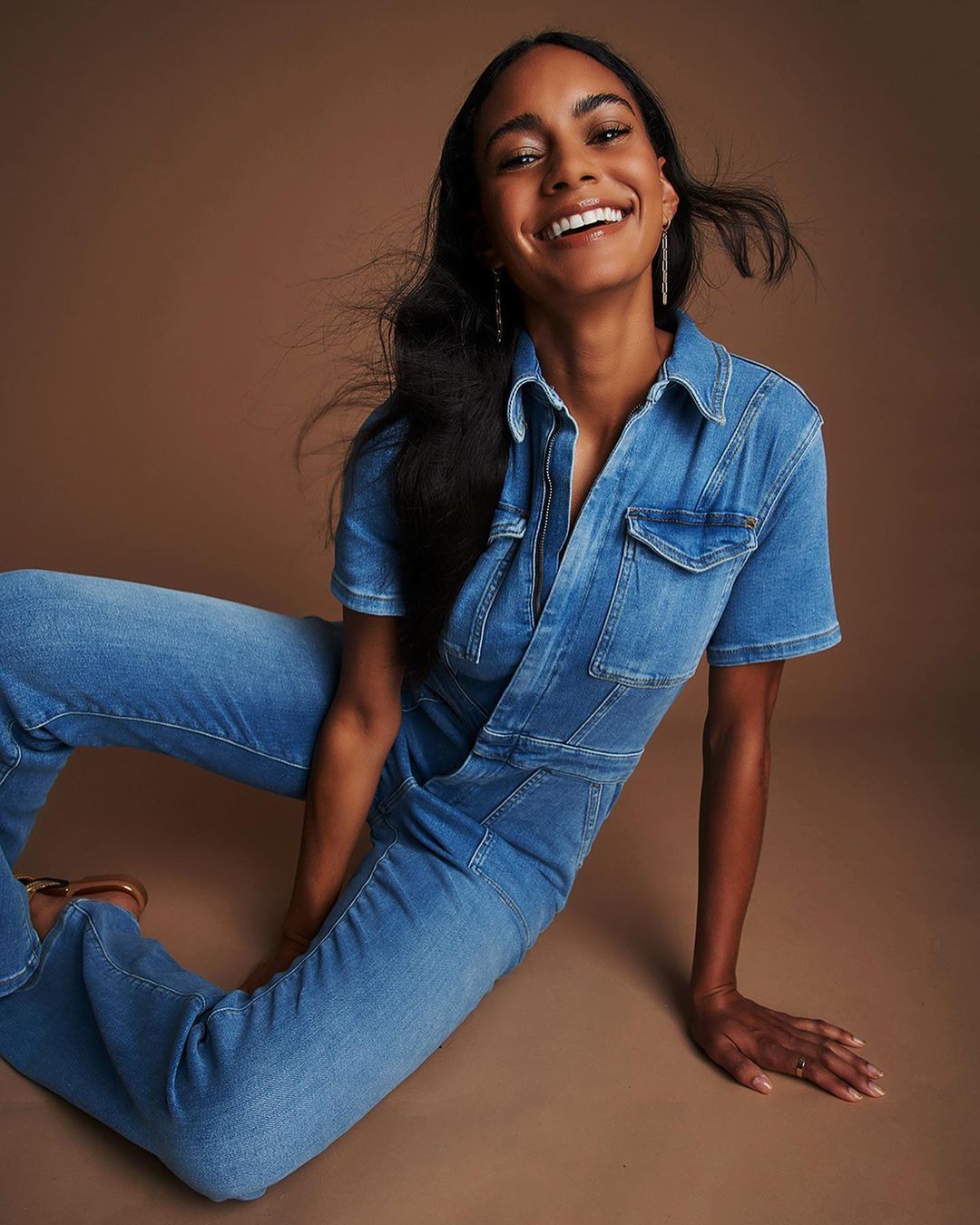 Smiling Black woman poses indoors wearing denim jumpsuit. She sits against a brown backdrop with light skin and dark hair.