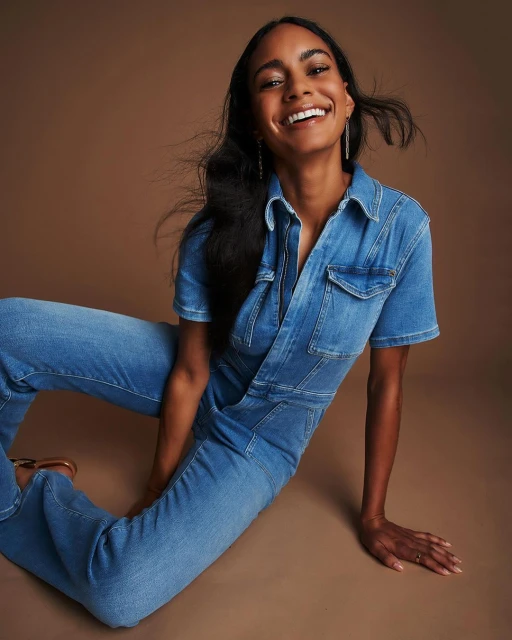 Smiling Black woman poses indoors wearing denim jumpsuit. She sits against a brown backdrop with light skin and dark hair.