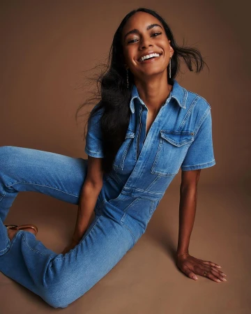 Smiling Black woman poses indoors wearing denim jumpsuit. She sits against a brown backdrop with light skin and dark hair.