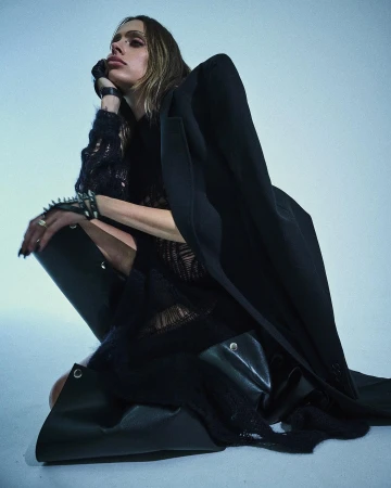 Woman in a black blazer, thigh-high boots, and a mesh top is kneeling in a photo studio with a neutral background, looking at the camera.