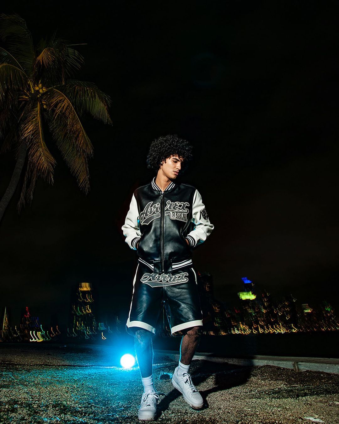 Stylish man poses in a black and white varsity jacket and shorts at night near a palm tree, with a city skyline in the distance.