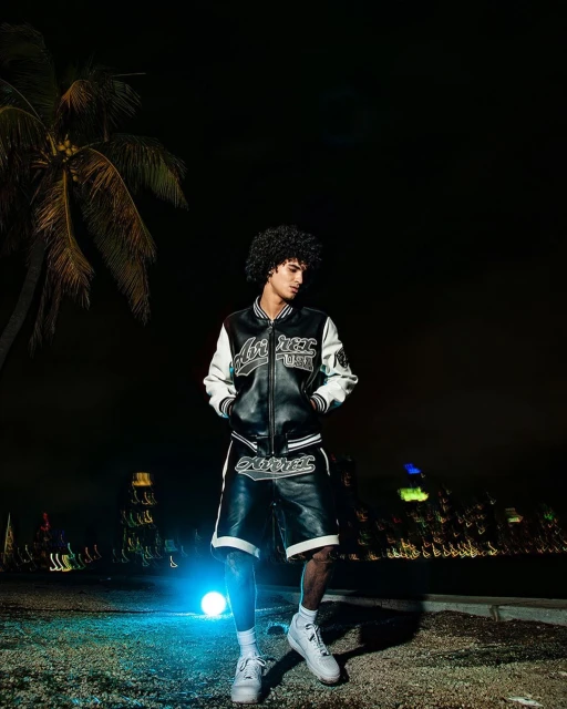 Stylish man poses in a black and white varsity jacket and shorts at night near a palm tree, with a city skyline in the distance.