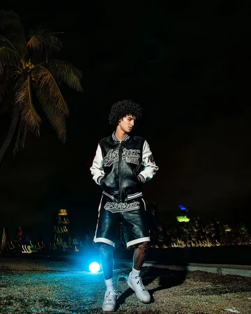 Stylish man poses in a black and white varsity jacket and shorts at night near a palm tree, with a city skyline in the distance.