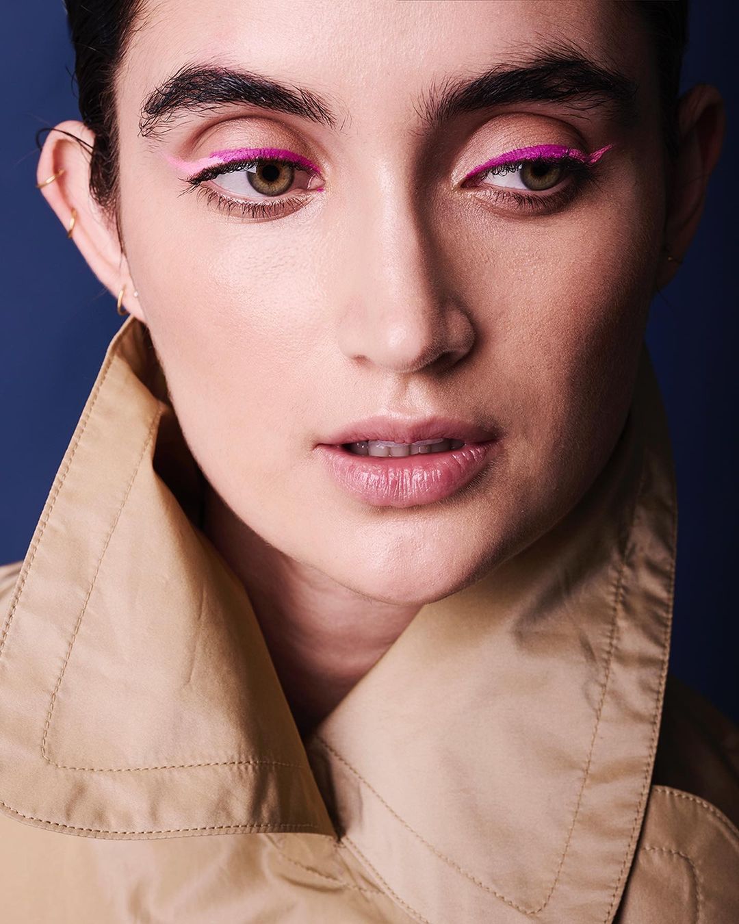 Close-up portrait of a woman with bright pink eyeliner wearing a tan jacket against a dark blue background in a beauty studio shot.
