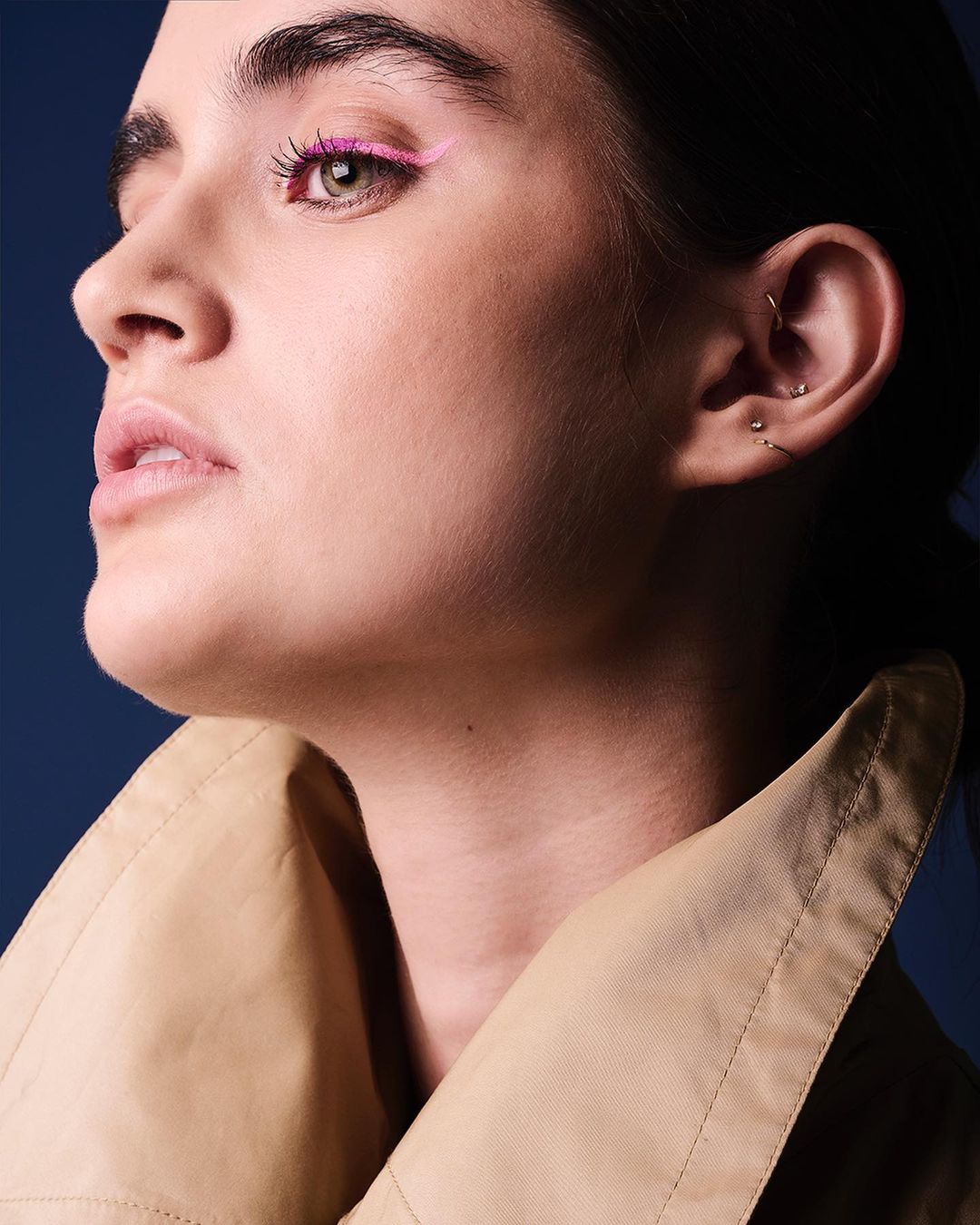 Close-up of a woman with pink eyeliner and a tan trench coat against a dark blue background. Beauty and fashion-themed image.