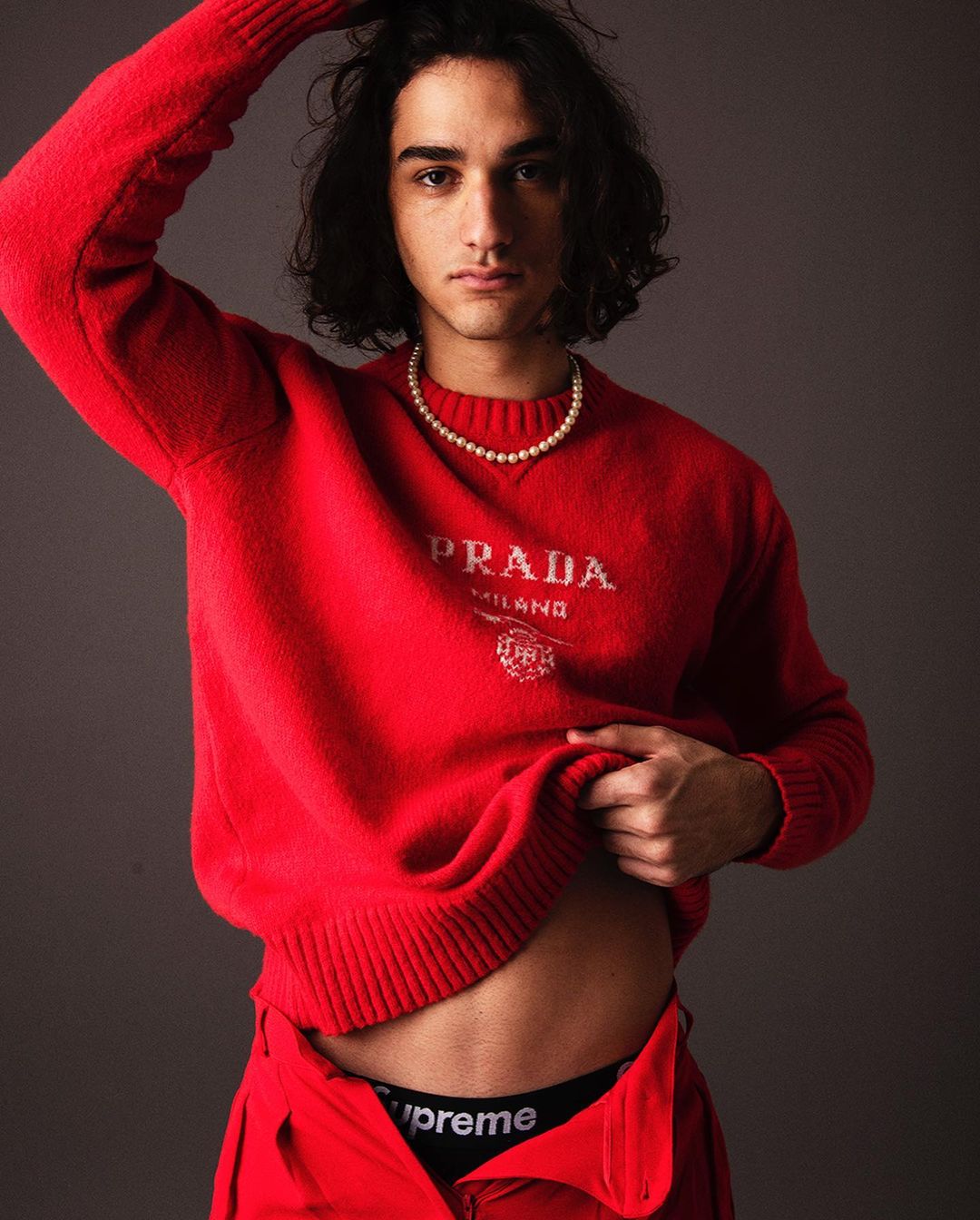 A young adult male poses indoors wearing a red Prada sweater and black Supreme-branded underwear, with a pearl necklace.