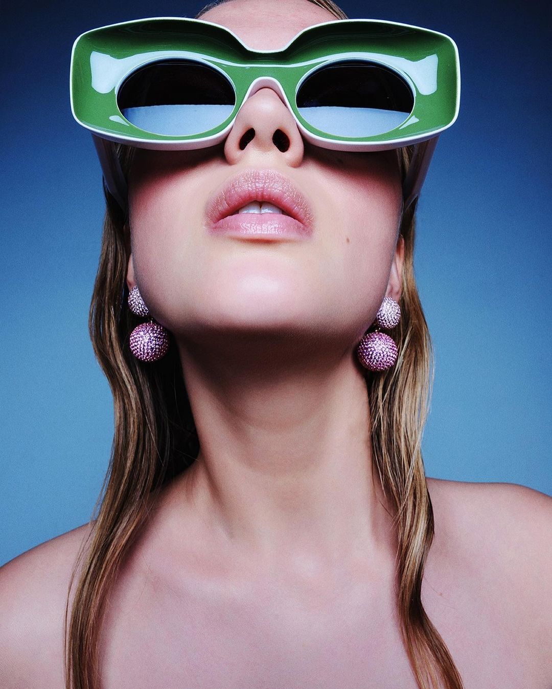Eye-level portrait of a woman wearing green-rimmed sunglasses and round pink earrings against a graduated blue background.