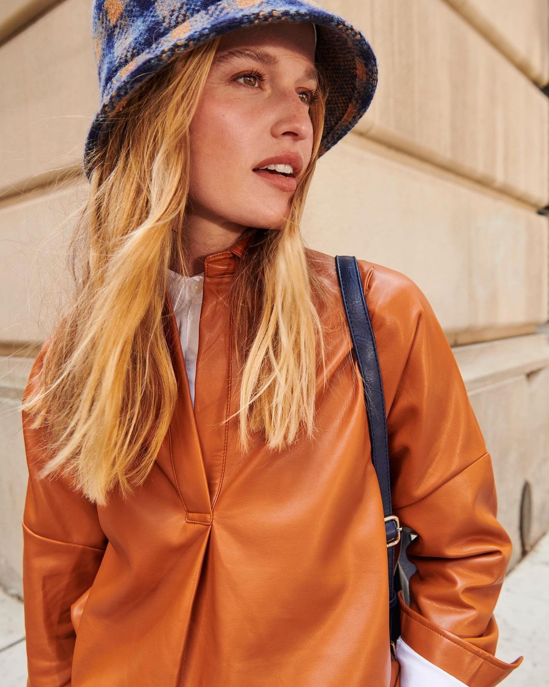 Portrait shot of a fashionable woman with long blonde hair wearing a rust-colored leather top and blue plaid bucket hat outdoors.