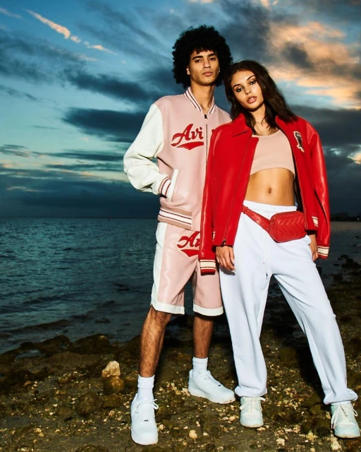 Two fashion models pose outdoors wearing matching pink varsity jackets and white athletic pants near the ocean at sunset.