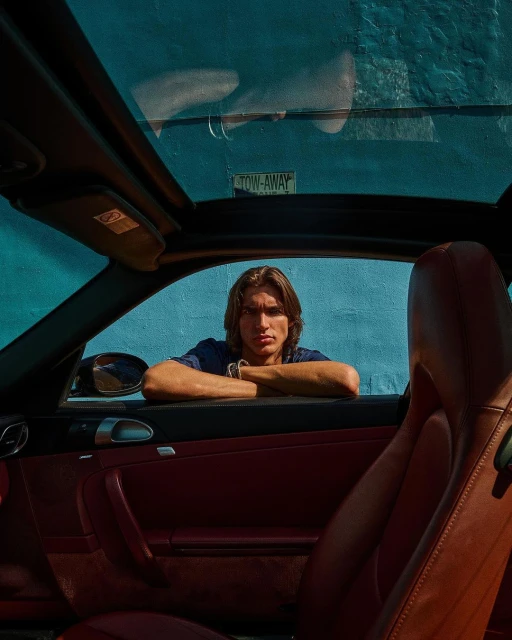 Adult male poses in a red car with a convertible top against a blue wall with a Tow-Away sign; interior shot with diffused light.