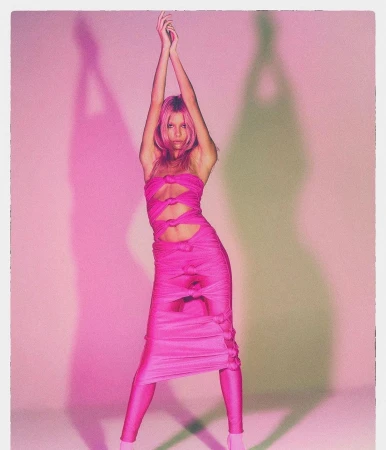 Full-length shot of adult woman in a pink dress with her arms raised above her head in a studio setting with pink lighting.