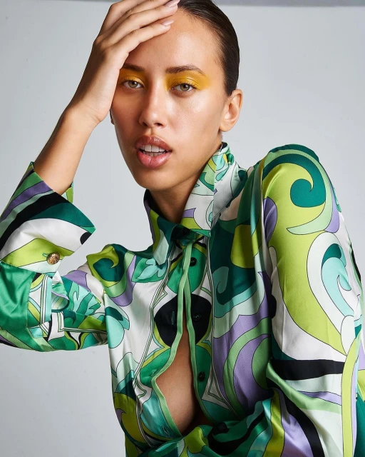 Fashion portrait of a woman with yellow eyeshadow, wearing an open green and purple swirl print blouse against a grey backdrop.