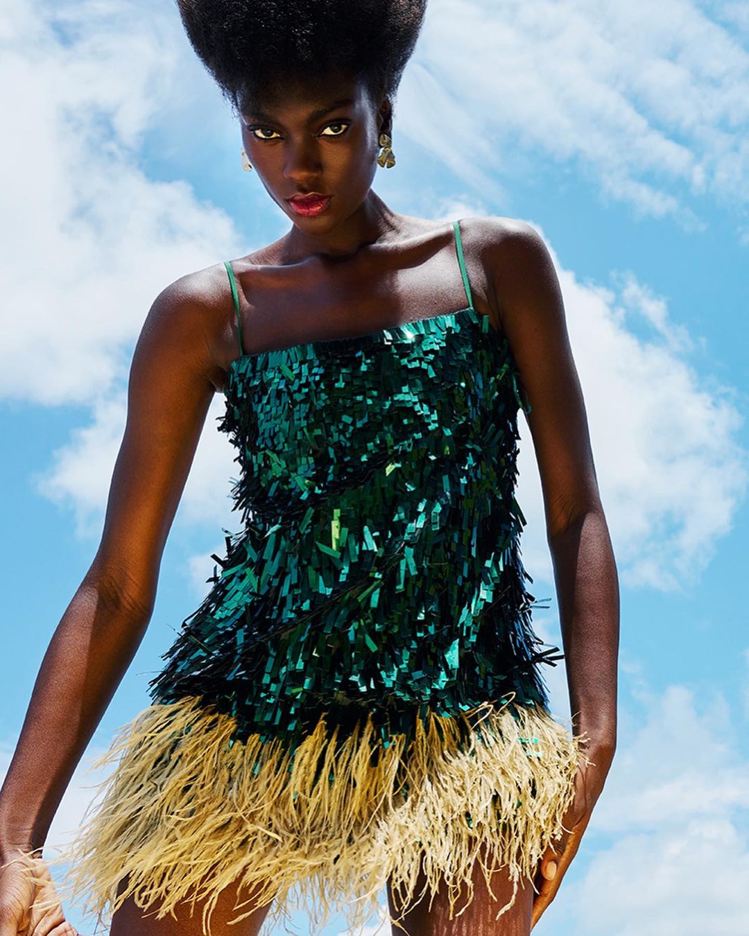 A young Black female model poses in a short, shimmering green dress with feathered hem against a bright blue sky filled with white clouds.