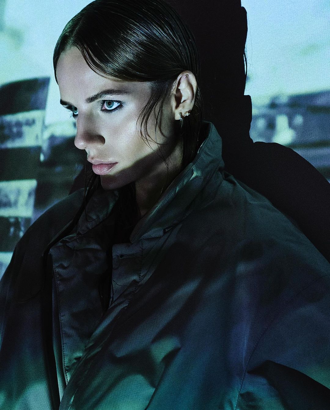 Portrait of a young adult woman in an olive-green jacket with dark hair and eyeliner, posing in front of a blue brick wall.
