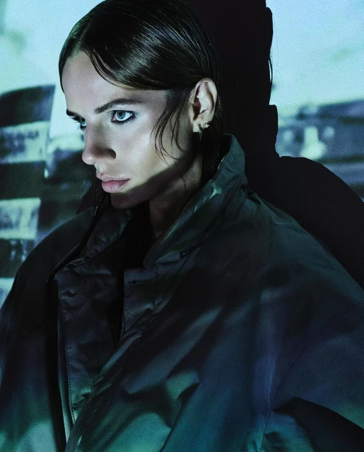 Portrait of a young adult woman in an olive-green jacket with dark hair and eyeliner, posing in front of a blue brick wall.