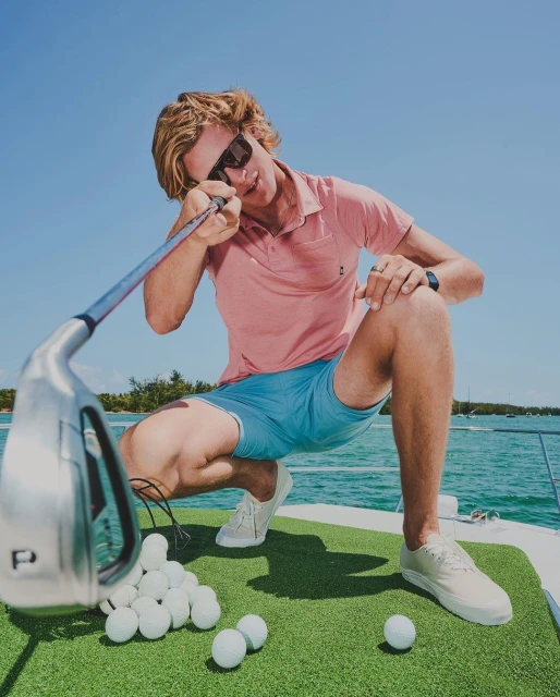 Man in sunglasses crouches on a boat with golf balls and golf club against a blue sky and water backdrop, outdoors.