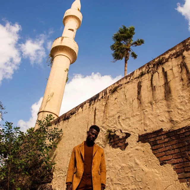 A man in a brown suit stands against a textured wall, with a tall tower, palm tree, and partly cloudy blue sky in the background.