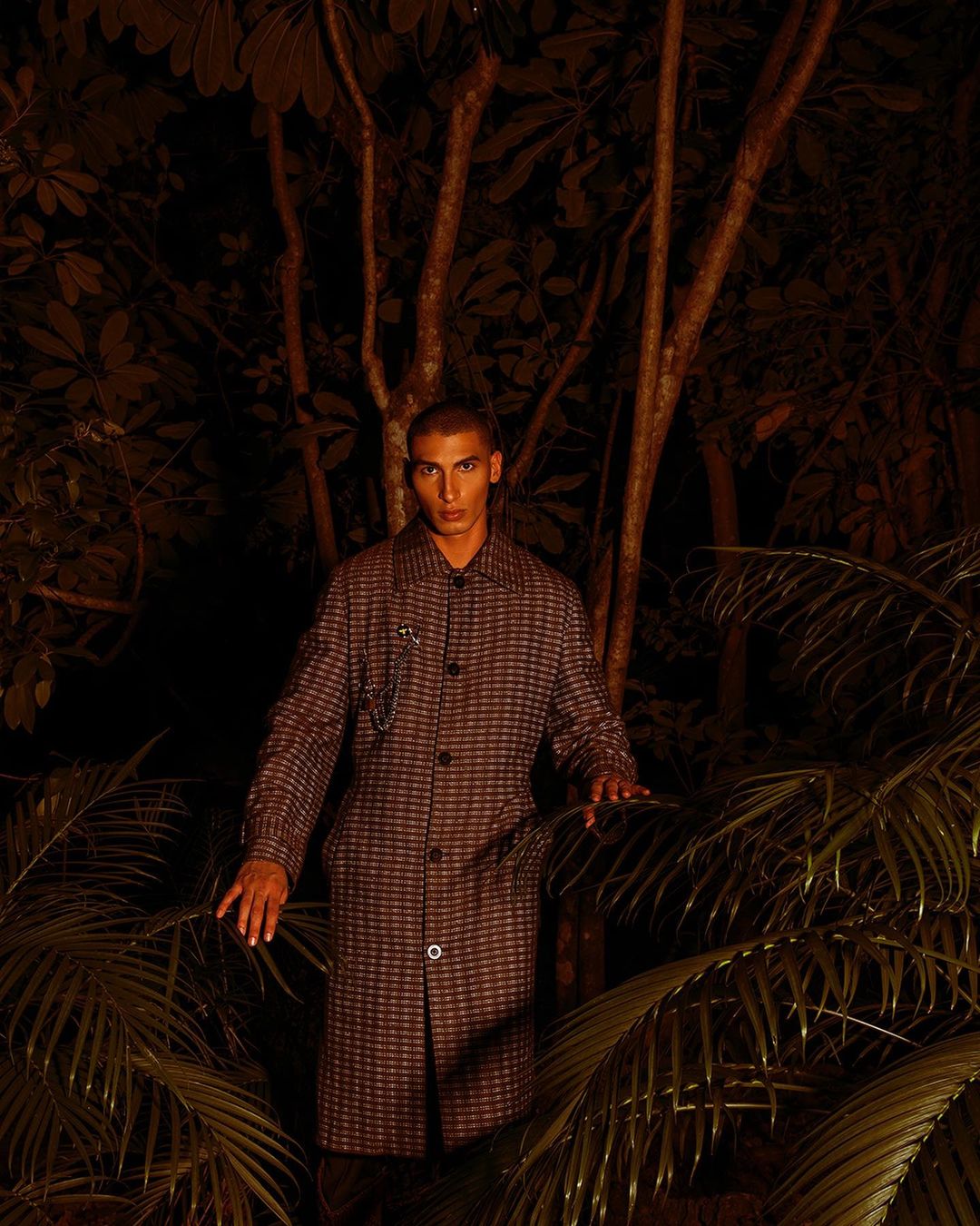 Adult male in a long, patterned coat poses amongst tropical foliage in a low-light, natural outdoor environment.