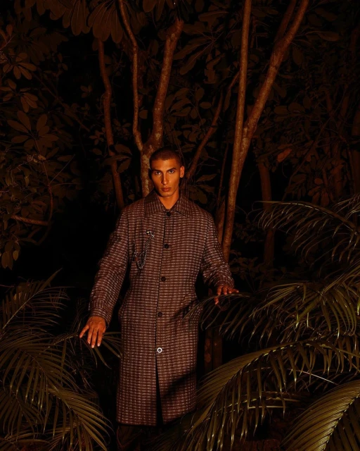 Adult male in a long, patterned coat poses amongst tropical foliage in a low-light, natural outdoor environment.