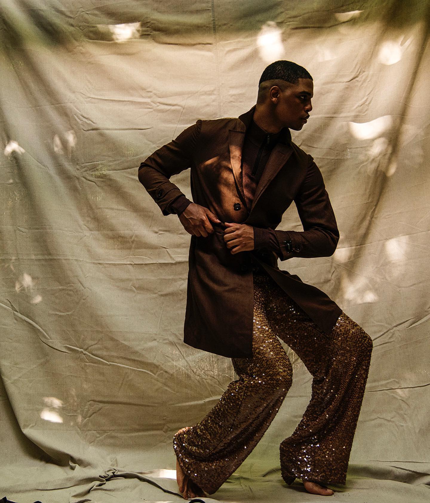 Fashionable black male model strikes a pose wearing a brown coat, shirt, and gold sequin pants against a textured backdrop.