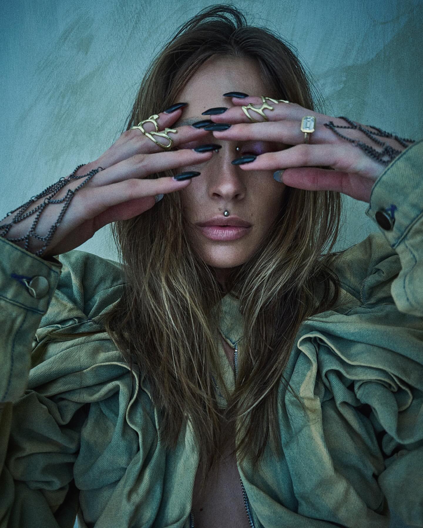 Close-up portrait of a woman with long dark blonde hair covering her eyes with hands featuring gold rings and long black nails.