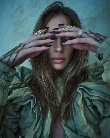 Close-up portrait of a woman with long dark blonde hair covering her eyes with hands featuring gold rings and long black nails.