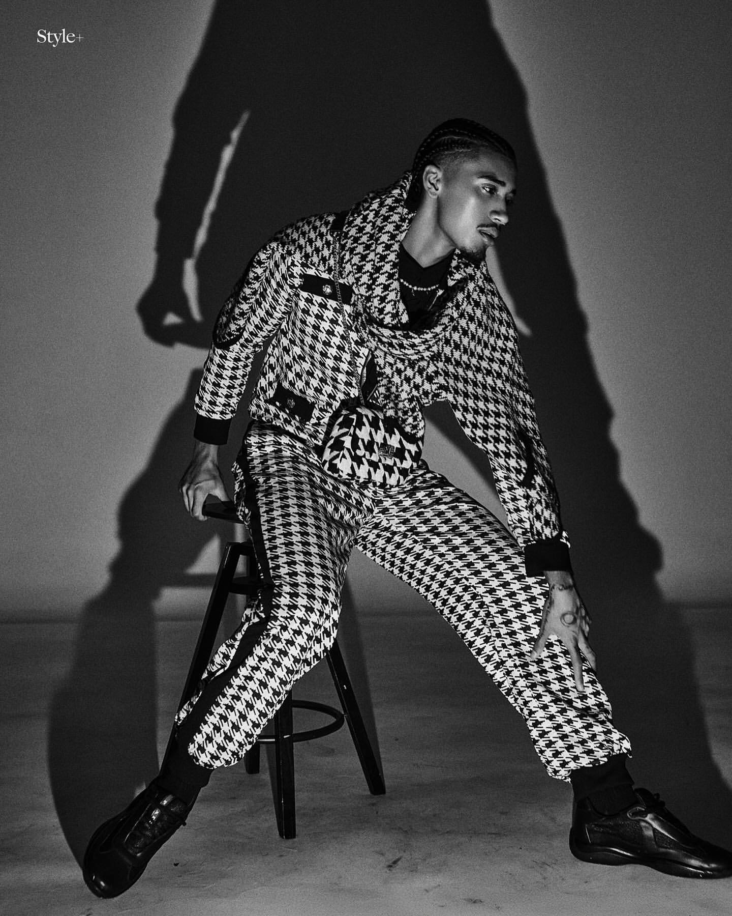 A man in a houndstooth suit sits on a stool against a wall with a shadow, showcasing fashion and style in a black-and-white photograph.