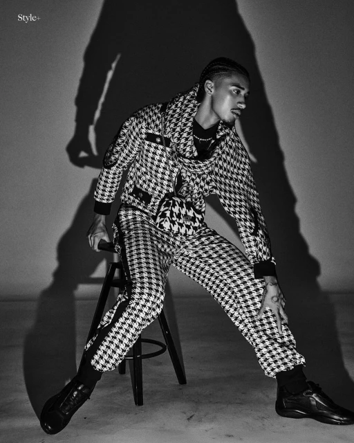 A man in a houndstooth suit sits on a stool against a wall with a shadow, showcasing fashion and style in a black-and-white photograph.