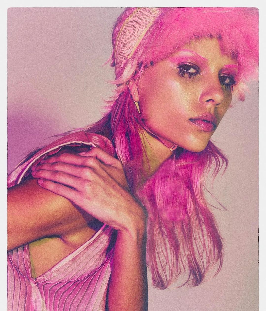 Close-up portrait of a woman with bright pink hair and dramatic makeup. She wears a pink top and a feathered hat in a photo studio.