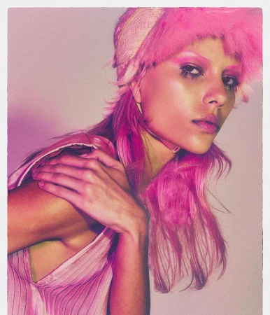 Close-up portrait of a woman with bright pink hair and dramatic makeup. She wears a pink top and a feathered hat in a photo studio.
