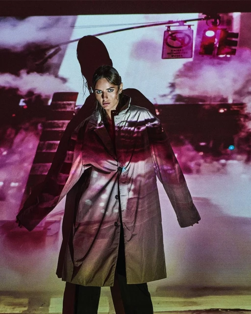 A woman in a long coat stands against a projected city background with streetlights, creating a visually striking, fashionable composition.