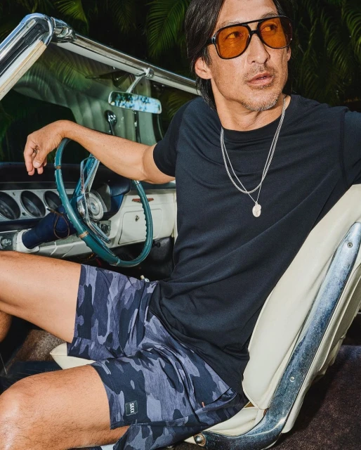 Man in sunglasses poses in a vintage convertible car wearing a black t-shirt and camouflage shorts against a palm tree backdrop at night.