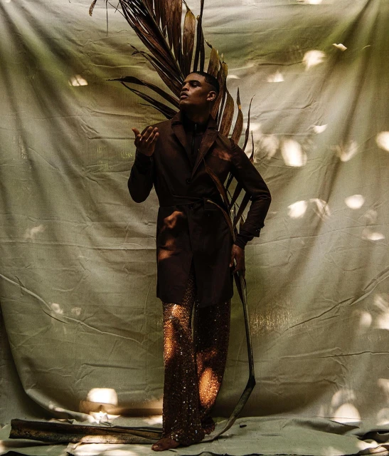 Fashionable man poses in a brown coat and sparkling pants, framed by dried palm fronds against a textured backdrop with soft, filtered light.