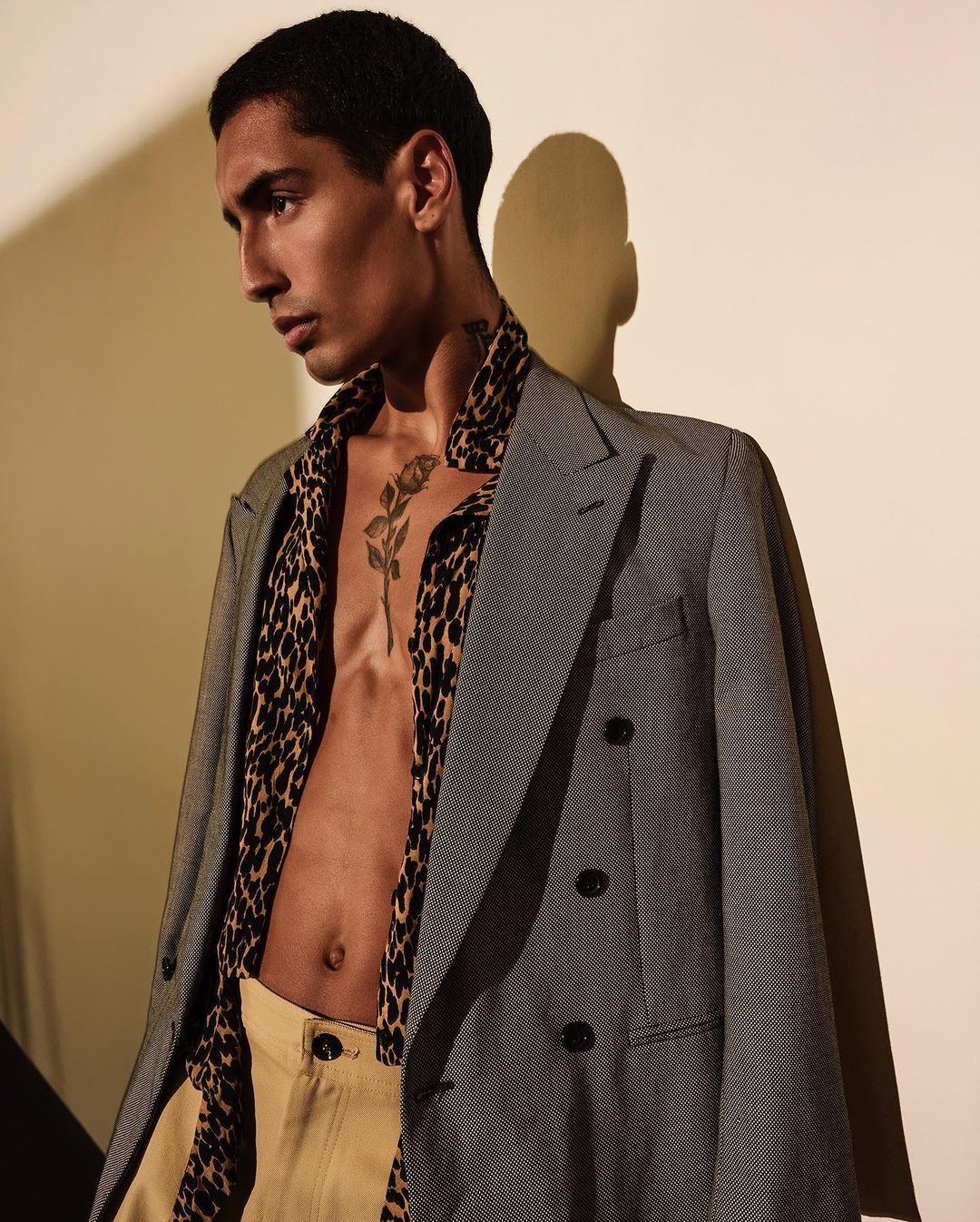 Attractive man with a tattoo poses indoors wearing a jacket over a partially unbuttoned animal-print shirt and khaki pants, eyes averted.