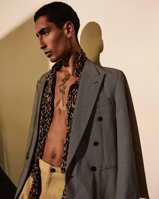 Attractive man with a tattoo poses indoors wearing a jacket over a partially unbuttoned animal-print shirt and khaki pants, eyes averted.