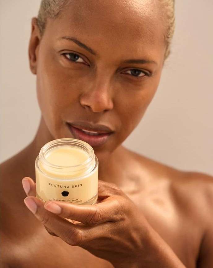 Close-up of an adult female with light skin holding a jar of Fortuna Skin cleansing balm, beauty and personal care product, in a neutral setting.