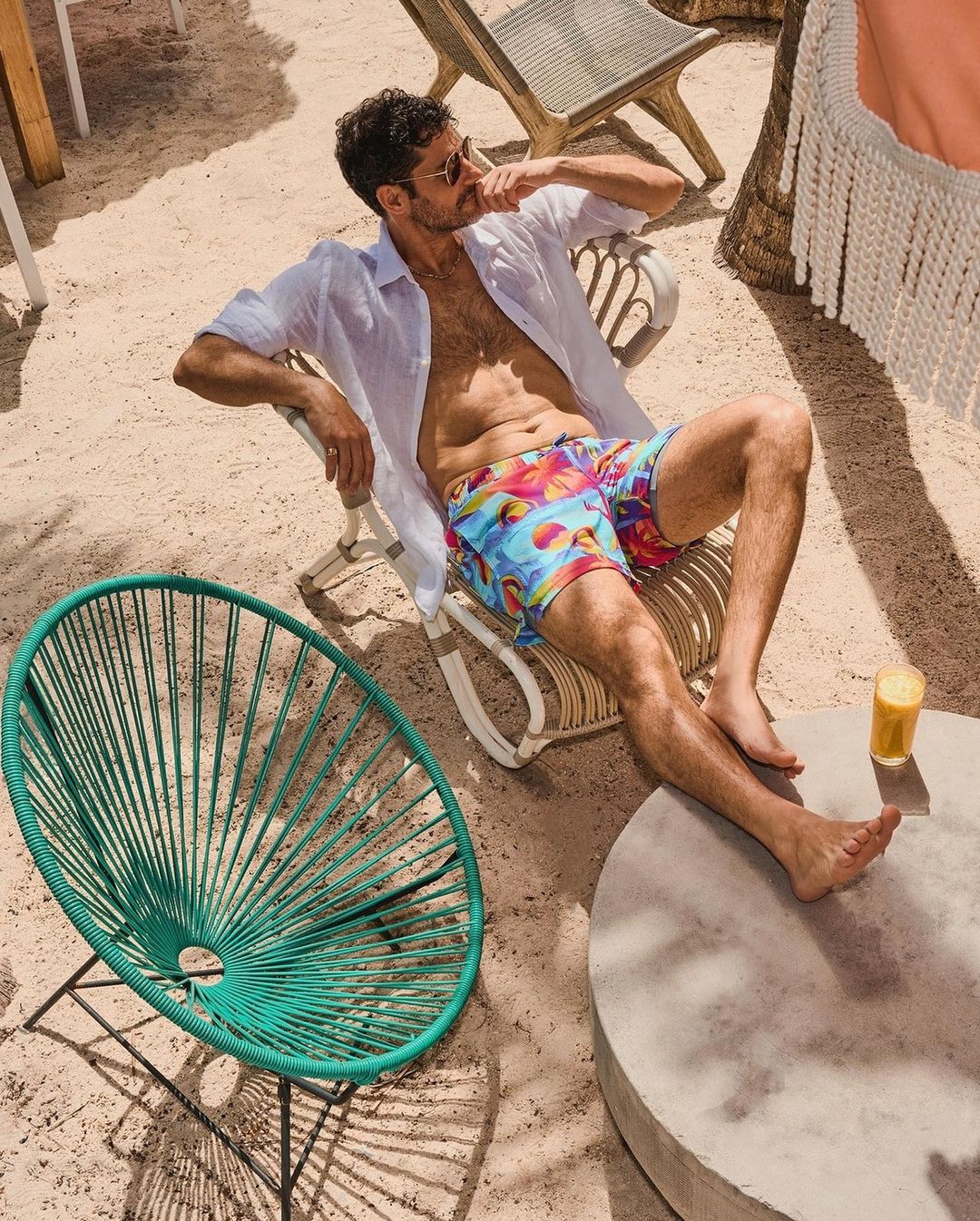 Man in swim trunks sits in a chair on a sandy beach with a drink, wearing an unbuttoned white shirt and sunglasses on a sunny day.