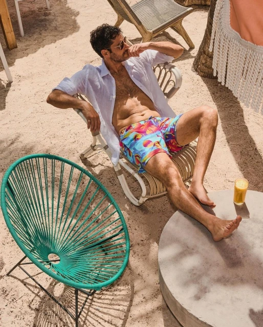 Man in swim trunks sits in a chair on a sandy beach with a drink, wearing an unbuttoned white shirt and sunglasses on a sunny day.