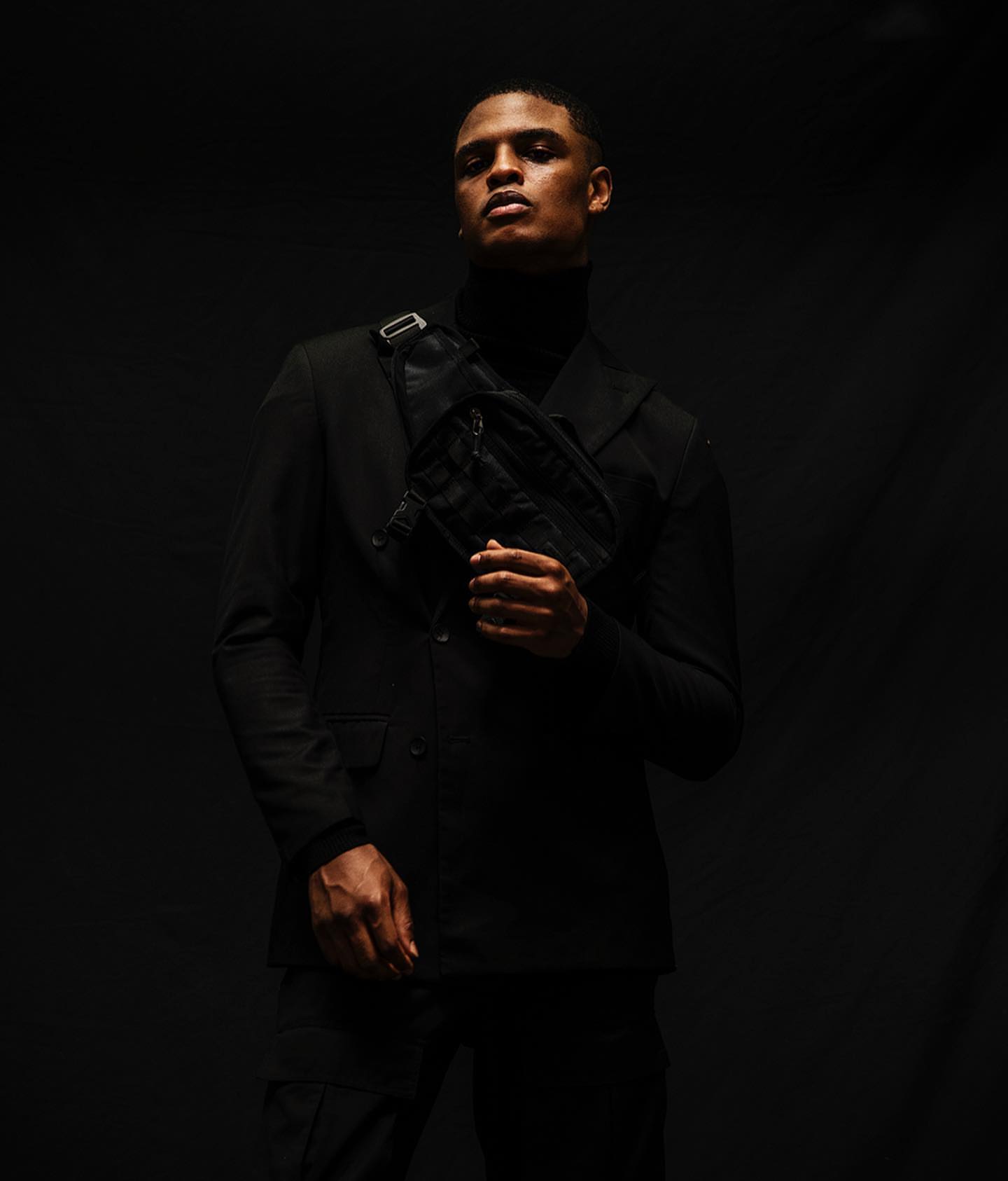 A stylish Black man poses in a black blazer and black turtleneck, accessorized with a modern black waist bag, against a dark backdrop.