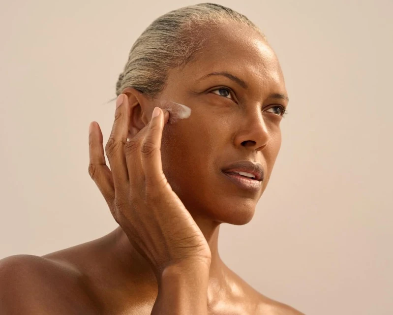 Close-up of an adult woman applying cream to her face. She has short gray hair and medium brown skin against a neutral background.