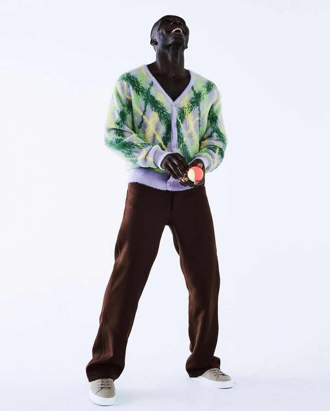An African male model in brown pants, a light green argyle sweater, and light-colored shoes poses on a white backdrop.