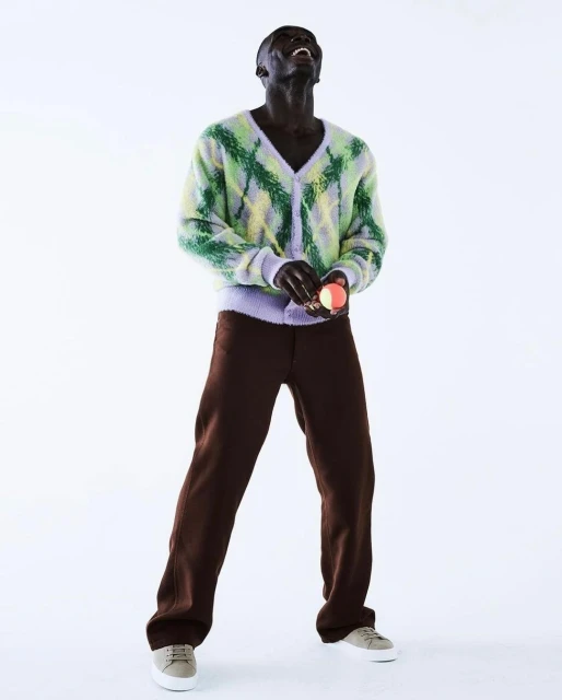 An African male model in brown pants, a light green argyle sweater, and light-colored shoes poses on a white backdrop.