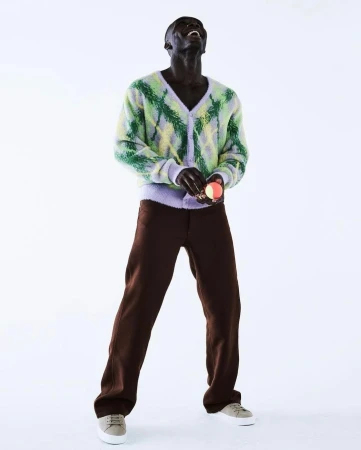 An African male model in brown pants, a light green argyle sweater, and light-colored shoes poses on a white backdrop.