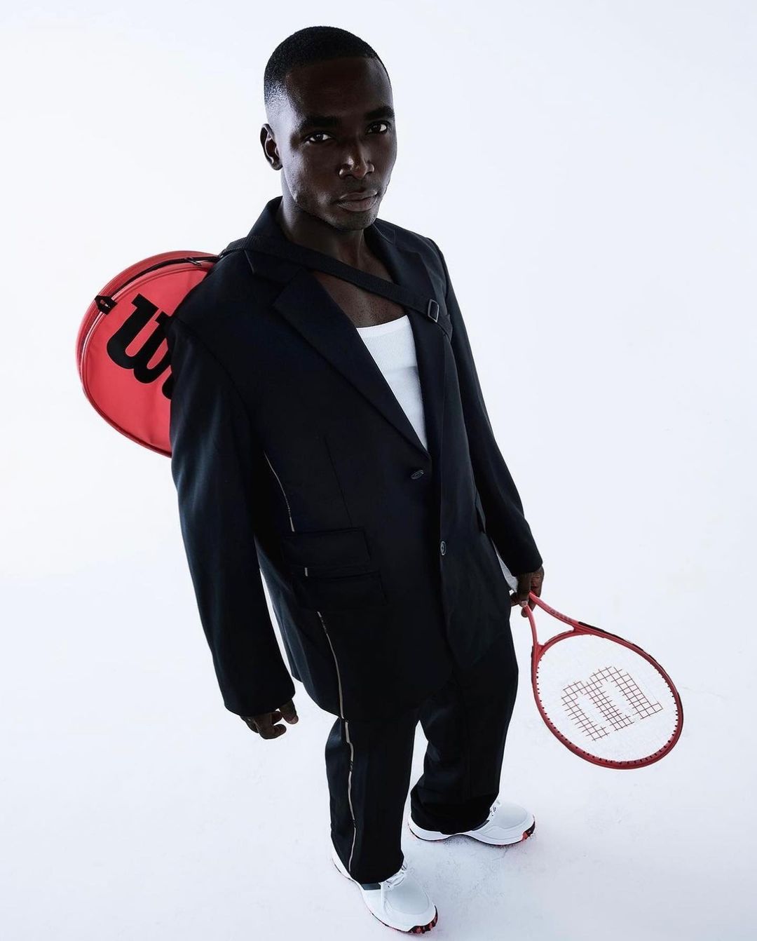 Man in suit posing with Wilson tennis racket and bag against a white backdrop in a high-angle studio shot. Fashion and sport concept.