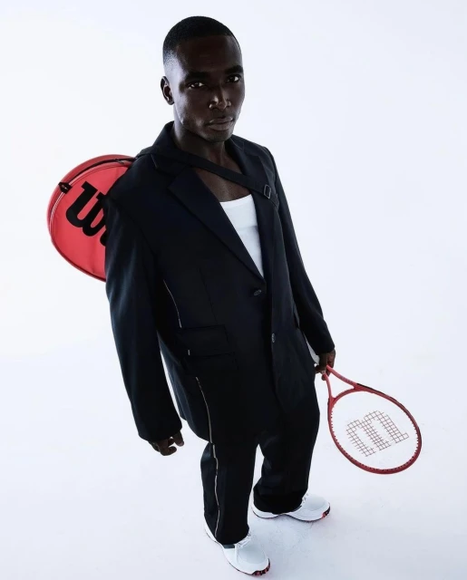 Man in suit posing with Wilson tennis racket and bag against a white backdrop in a high-angle studio shot. Fashion and sport concept.