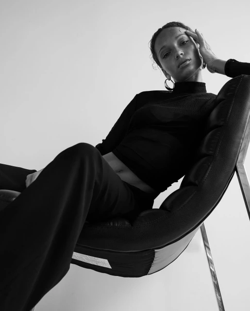 A woman in black attire poses while seated in a modern chair, captured from a low angle in a high contrast black and white studio setting.