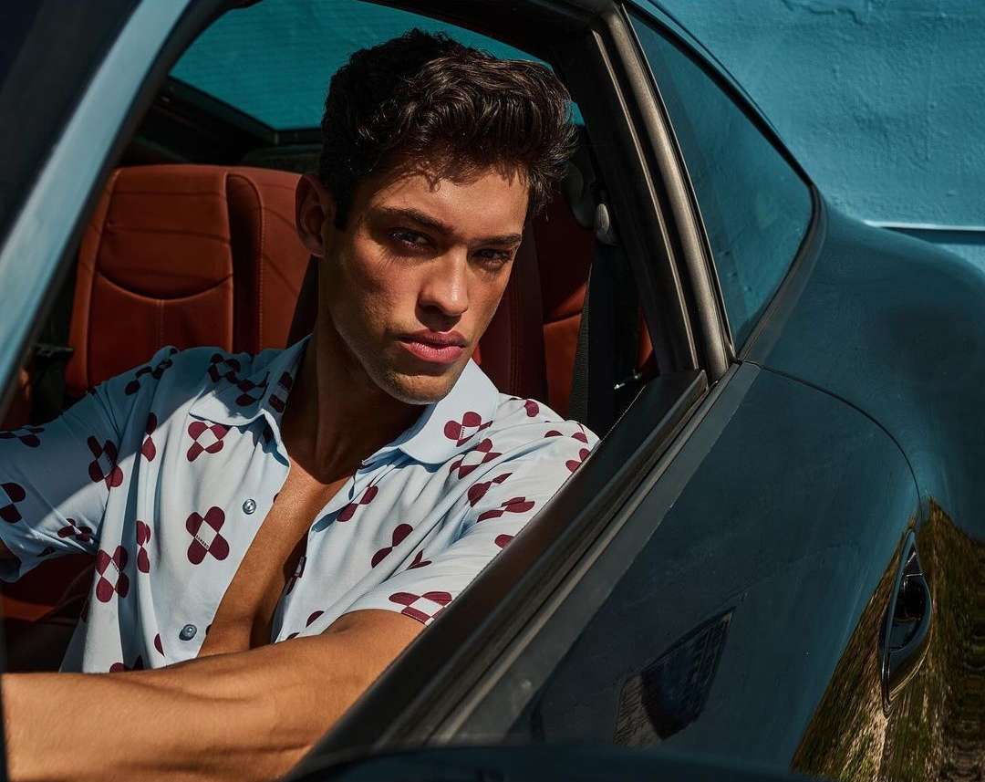 A young man with dark hair sits in a car, wearing a light blue floral shirt, posing for a fashion shot with a confident expression.