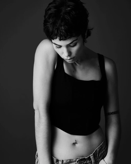 Monochrome portrait of a young woman with a pixie cut, black tank top, and jeans against a plain background looking down with arms relaxed.
