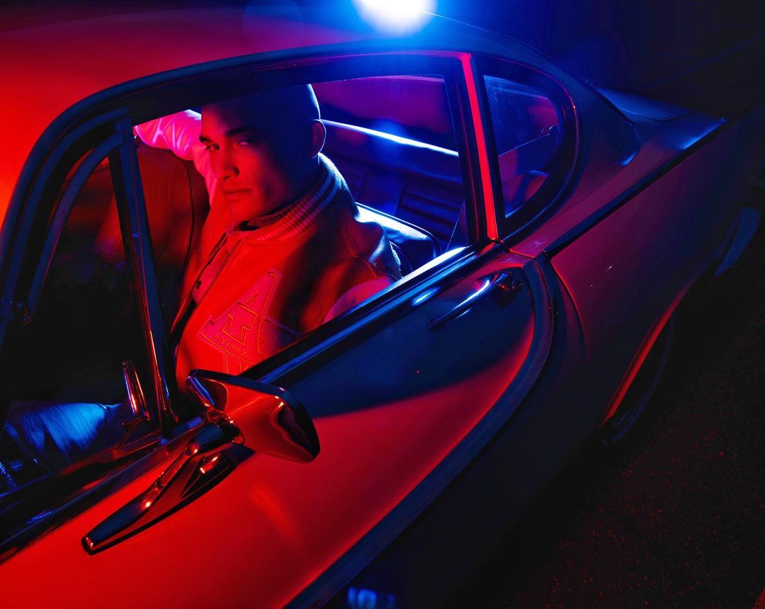 Asian man in a letterman jacket sits in the driver's seat of a vintage car, bathed in red and blue light for an automotive editorial.