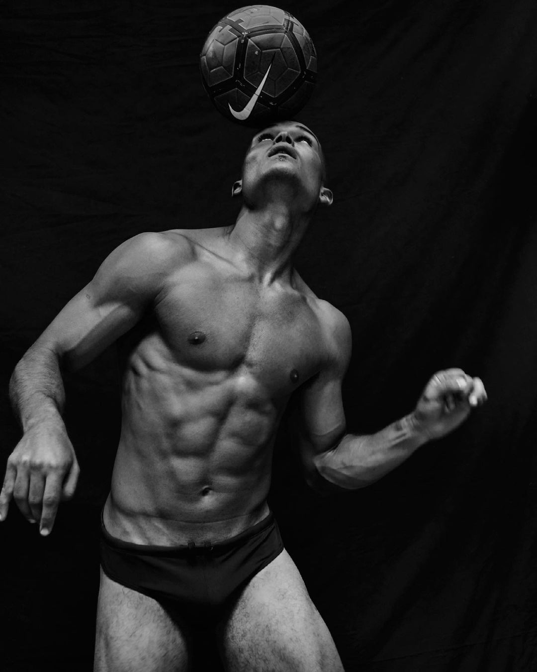 Shirtless man balances soccer ball on head against dark background. Athletic build is highlighted by soft lighting in this studio shot.